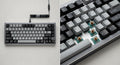 Clavier mécanique compact avec châssis en métal argenté, touches PBT grises et noires, switches lubrifiés visibles sous certaines touches.